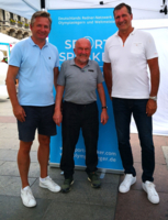 Olympiasieger Klaus Wolfermann und zwei weitere Herren stehen vor einem Plakat.