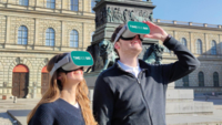 G&auml;ste des TimeRide tragen eine VR Brille vor der Residenz in M&uuml;nchen.