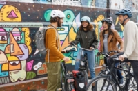 Vier Fahradfaher*innen mit Helm stehen vor einer mit Graffiti bemalten Wand.