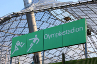 Gr&uuml;nes Hinweisschild im Olympiastadion M&uuml;nchen, Zeltdach im Hintergrund