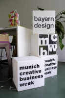 Auf wei&szlig;en, &uuml;bereinander gestapelten Leucht-W&uuml;rfeln, steht in schwarzer Schrift "bayern design", "Munich Creative Business Week" und "mcbw" geschrieben.