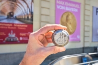 A close-up of a hand holding a large silver coin bearing the image of Munich&rsquo;s Frauenkirche.