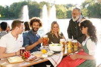A group of humans is sitting on benches in a beer garden in Munich. On a table between them is traditional food and beer.