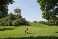 Zwei Frauen gehen auf der Wiese unter dem Monopteros in M&uuml;nchen.