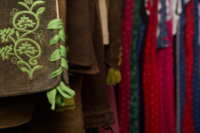 Nahaufnahme einer bestickten Lederhose mit verschiedenen Dirndl im Hintergrund.