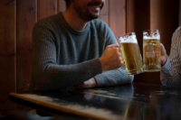 Two people at a wooden table in a pub in Munich toast each other with two glass beer mugs.