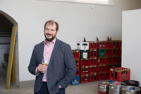 Ein Mann steht mit einem Bierglas in der Hand vor vielen Bierk&auml;sten und F&auml;ssern.