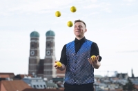 A man wearing a black-and-white waistcoat, black trousers and a shirt is juggling yellow balls against the backdrop of Munich&rsquo;s Frauenkirche.
