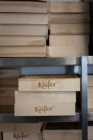 Holzboxen mit dem K&auml;fer Logo liegen gestapelt in einem Regal.