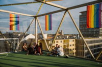 Several people sit on the roof terrace of Bellevue di Monaco in Munich and rainbow flags hang on the building.