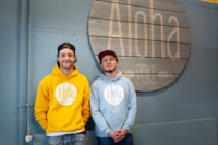 Die beiden Gr&uuml;nder, junge M&auml;nner in gelben und blauen Hoody und Basecap vor dem Schild &bdquo;Aloha Poke&ldquo; Ihres Ladens.