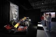 A couple of teenagers are taking photos of a racing car as part of the Formula 1 exhibition; behind it hangs a black-and-white portrait of the driver.
