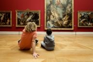 A young man and a boy are sitting on the ground in front of paintings in the Alte Pinakothek in Munich.