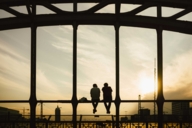 Zwei Personen sitzen auf dem Geländer der Hackerbrücke und schauen in Richtung Sonnenuntergang.