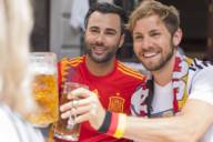 Zwei junge Männer, einer davon im Trikot der spanischen, der andere in dem der deutschen Nationalmannschaft, prosten einer dritten Person zu.