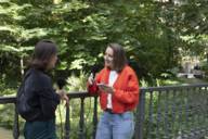Zwei Frauen stehen mit Mikrofonen auf einer Brücke im Grünen und unterhalten sich.