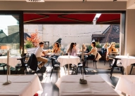 Vue de l'int&eacute;rieur vers l'ext&eacute;rieur sur la terrasse sur le toit de l'Anagram - Haus im Tal, o&ugrave; se trouvent quelques petites tables rondes autour desquelles des personnes sont assises et d&eacute;gustent des boissons.