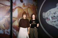 Fotografie von zwei Frauen, die vor einem Kunstwerk stehen, auf dem links aufgerollte Schinkenscheiben und rechts die gezeichnete Schnauze eines Wildschweins zu sehen sind..