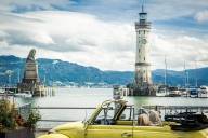 The harbour at Lindau with the Bavarian lion and the lighthouse; in the foreground, a yellow classic car; in the background, the mountains.