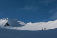 Zwei Skitourengänger auf dem Weg zum Gipfel vor blauem Himmel.