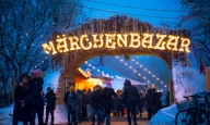 Ein Holztorbogen auf dem beleuchtet "M&auml;rchenbazar" steht und unter dem Menschen, die winterlich angezogen sind, hindurch gehen.