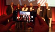 Ein Smartphone fotografiert Menschen auf einer Bühne beim Tourismustag in München