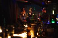 Eine Sängerin, ein Gitarrist und ein Cello-Spieler machen Musik in einer Bar in München.