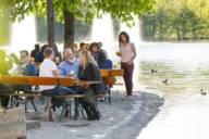 Eine Frau steht an einem Tisch in einem Biergarten in München. Im Hintergrund ist ein See.