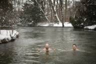 Zwei Männer schwimmen im Winter im Eisbach in München. Am Ufer liegt Schnee.
