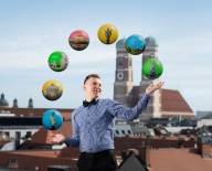 Ein Mann mit blauem Hemd, schwarzer Hose und Fliege jongliert Kugeln, die die Münchner Stadtviertel repräsentieren sollen. Im Hintergrund sieht man die Frauenkirche.