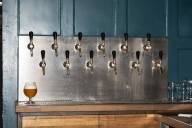 Eine Wand mit zwei Reihen von Zapfhähnen und einem eingeschenkten Bier in bauchigem Glas in der Bar BrewsLi.