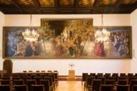 Blick auf ein großes Gemälde in einem Sitzungssaal im Neuen Rathaus in München. Davor hängen zwei Kronleuchter.