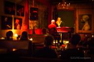 Blick in eine schummrig-beleuchtete Bar, im hinteren Teil eine kleine Bühne, wo ein Mann am Piano sitzt und eine Frau singt.