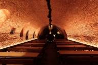In a large vaulted brick cellar, several tables and beer benches are lined up, and a number of wooden barrels are stacked up