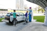 Ein Mann und eine Frau stehen mit einem E-Auto an einer Tankstelle an der BMW-Welt in München.