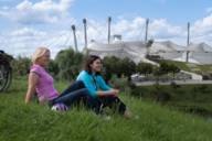 Zwei Frauen in bunter Kleidung sitzen auf dem Olympiaberg in München im Sommer. Im Hintergrund Fahrräder und das Olympiastadion.