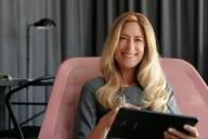 Eine Frau sitzt in einem Sessel und schaut lächelnd in die Kamera während sie ein Tablet in der Hand hält.