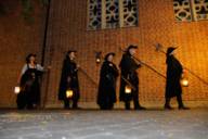 Mehrere Nachtwächter stehen mit Ihren Laternen vor einer Mauer.