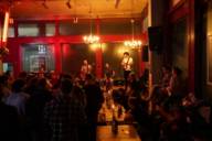 Eine Band aus drei Männern spielt Musik in einer gemütlichen Bar vor einem Publikum.