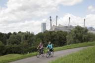 Zwei Frauen fahren auf dem Fahrrad auf einem Weg am Olympiaberg in München. Im Hintergrund das Olympiastadion.