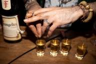 Vier Schnapsgläser gefüllt mit Frangelico Likör, der gold schimmert, stehen auf einem Holztisch, darüber sind zwei Hände zu sehen mit silbernen Ringen die eine Zigarette halten.