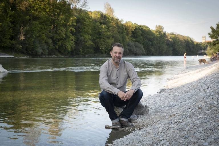 Flo Schairer, der Host des einfach München-Podcasts sitzt auf einem Stein am Kiesstrand der Isar in München.