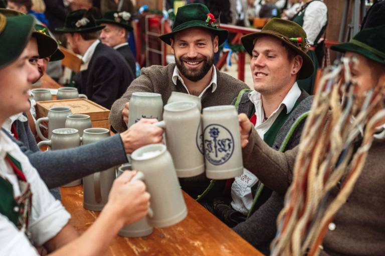 At a Wiesn regulars' table, several men in traditional costume toast each other with beer from traditional stoneware mugs.