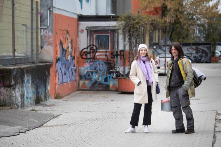 Eine Frau im Mantel und ein Künstler mit Farbeimer lächeln in die Kamera in München.
