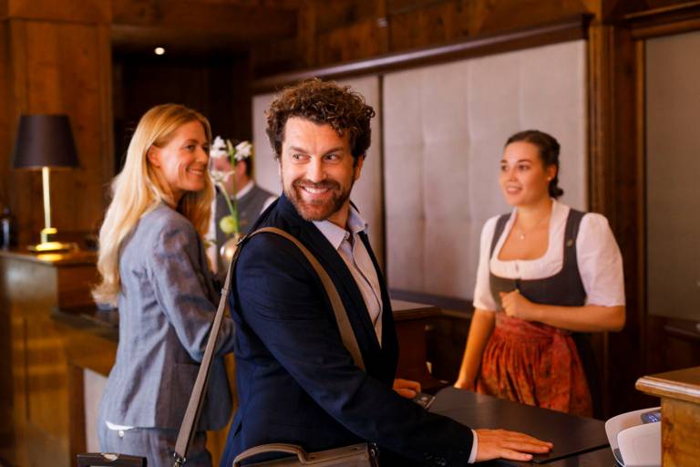 A woman dressed in a dirndl is welcoming a woman and a man in a hotel in Munich.