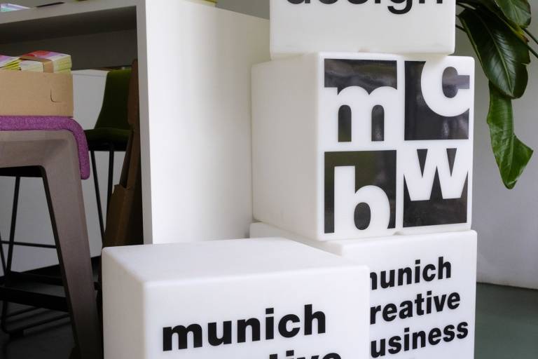 Auf weißen, übereinander gestapelten Leucht-Würfeln, steht in schwarzer Schrift "bayern design", "Munich Creative Business Week" und "mcbw" geschrieben.