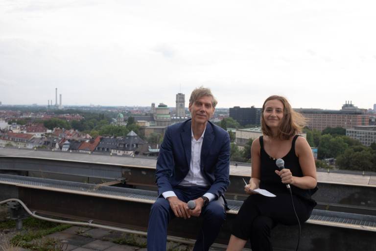 Ein Mann und eine Frau sitzen mit Mikrofonen auf einem Dach in München.