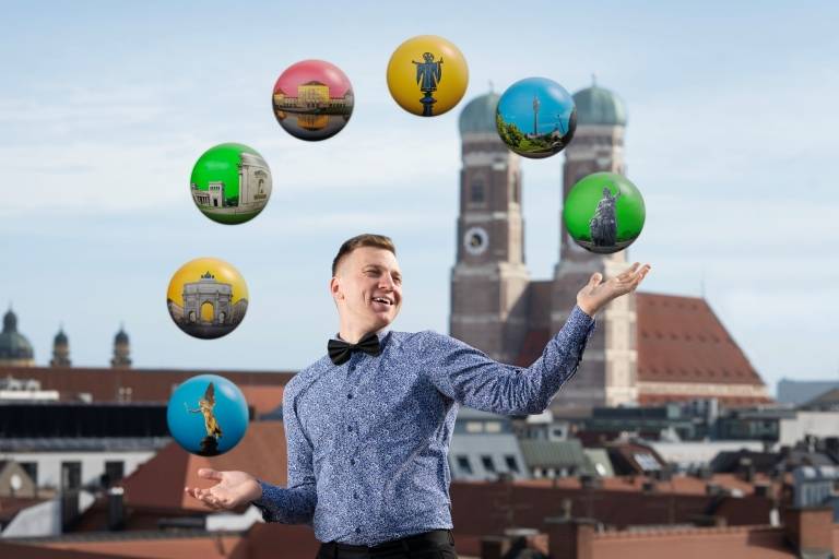 Ein Mann mit blauem Hemd, schwarzer Hose und Fliege jongliert Kugeln, die die Münchner Stadtviertel repräsentieren sollen. Im Hintergrund sieht man die Frauenkirche.