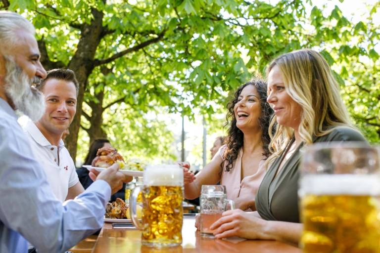 Zwei Männer und zwei Frauen sitzen an einem Tisch in einem Biergarten in München und prosten sich zu.