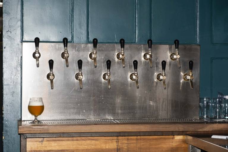 Eine Wand mit zwei Reihen von Zapfhähnen und einem eingeschenkten Bier in bauchigem Glas in der Bar BrewsLi.